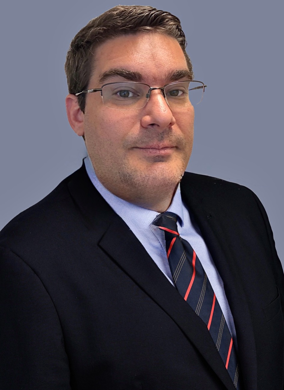 Professional headshot of a confident businessman in a dark suit, blue shirt, and striped tie, wearing glasses, posed against a neutral background.