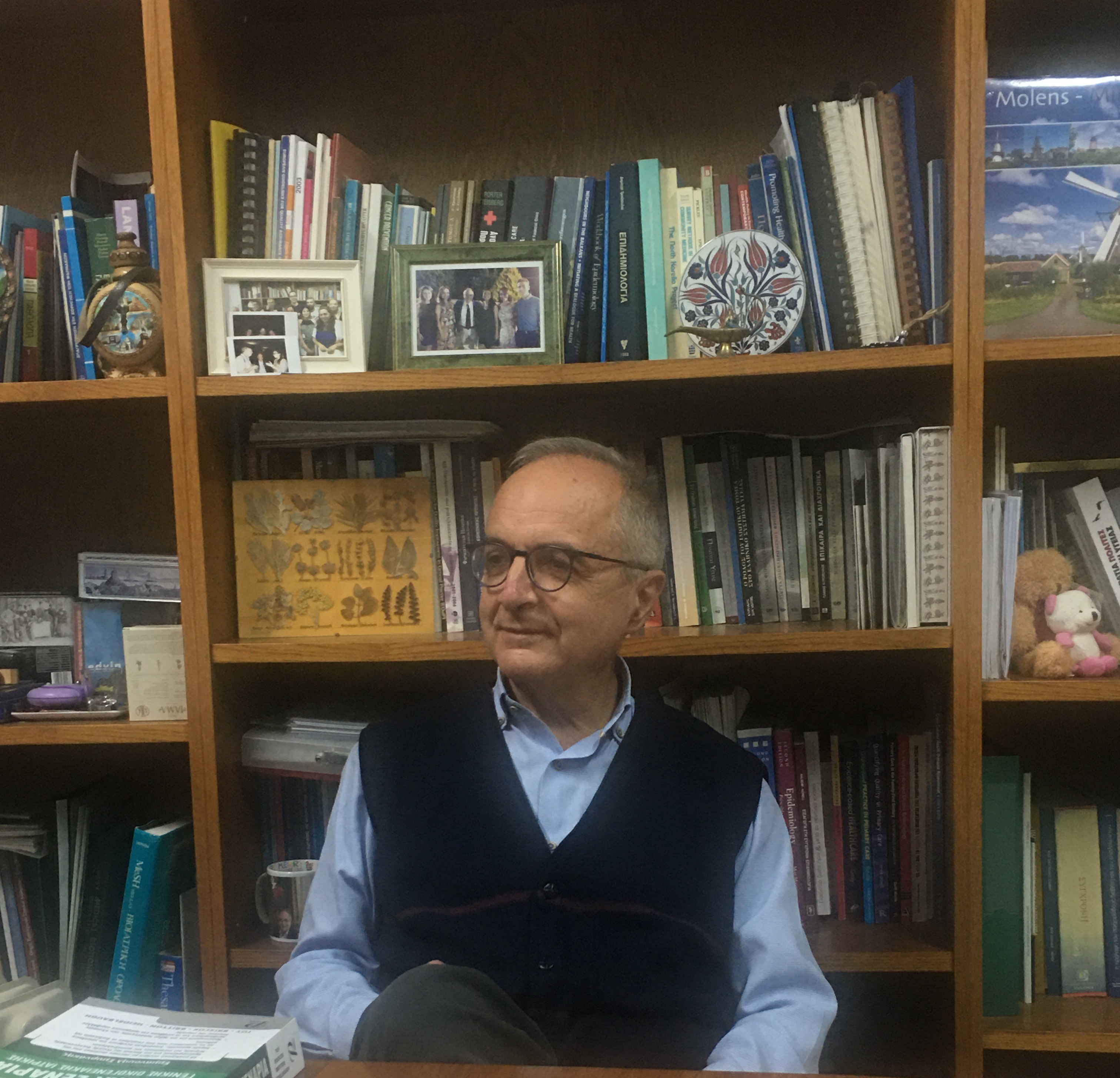 Portrait of a person wearing glasses and a dark vest over a light blue shirt, seated in front of a bookshelf filled with books, framed photographs, and decorative items in a study or office setting.
