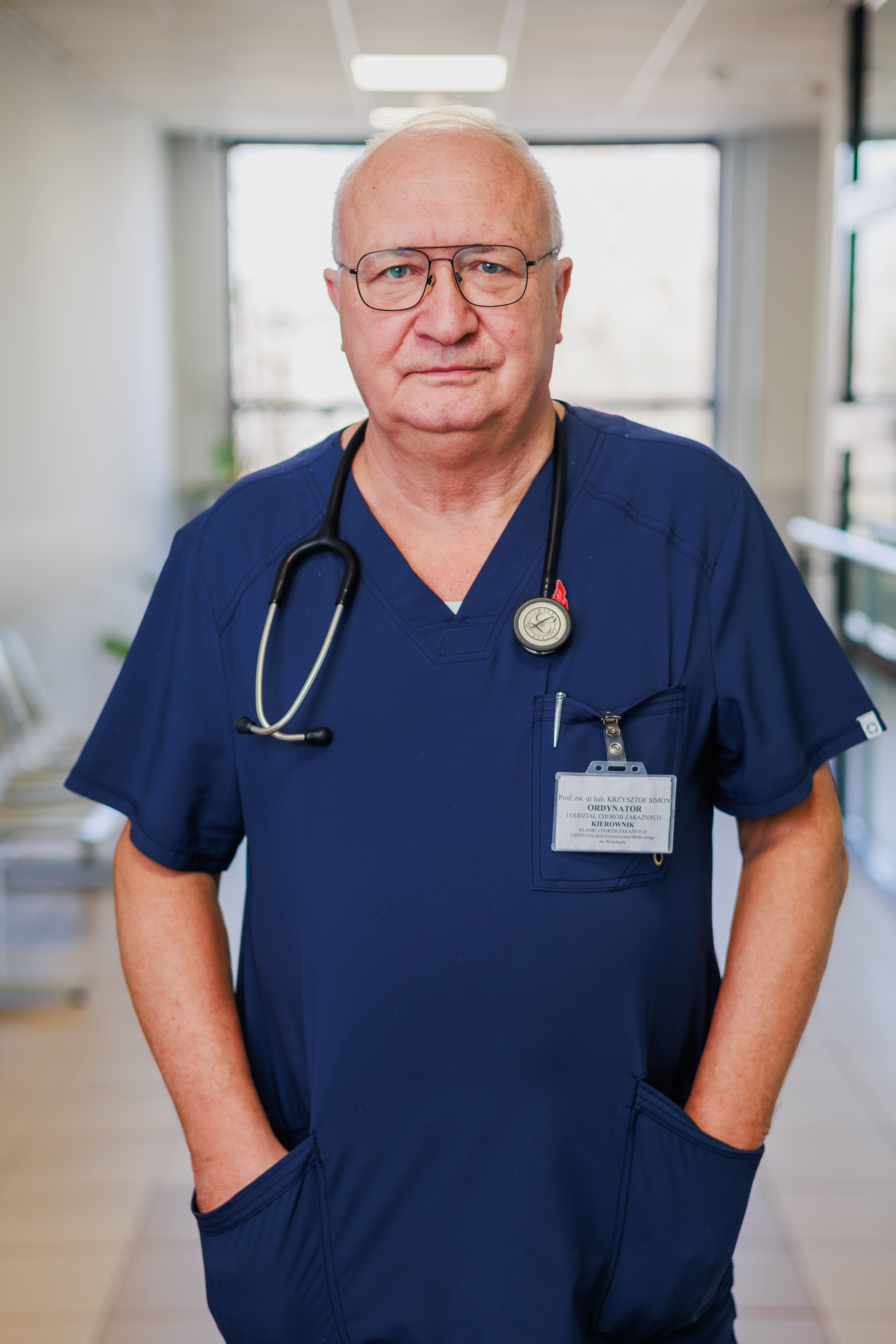 A male healthcare professional wearing navy blue medical scrubs and a stethoscope, standing in a bright hospital corridor.