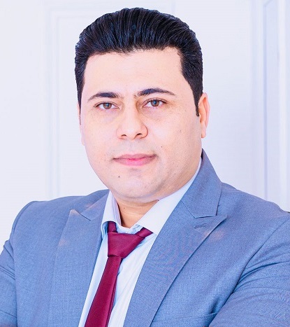 Portrait of a professional man in a light blue suit, white shirt, and red tie against a white paneled background.