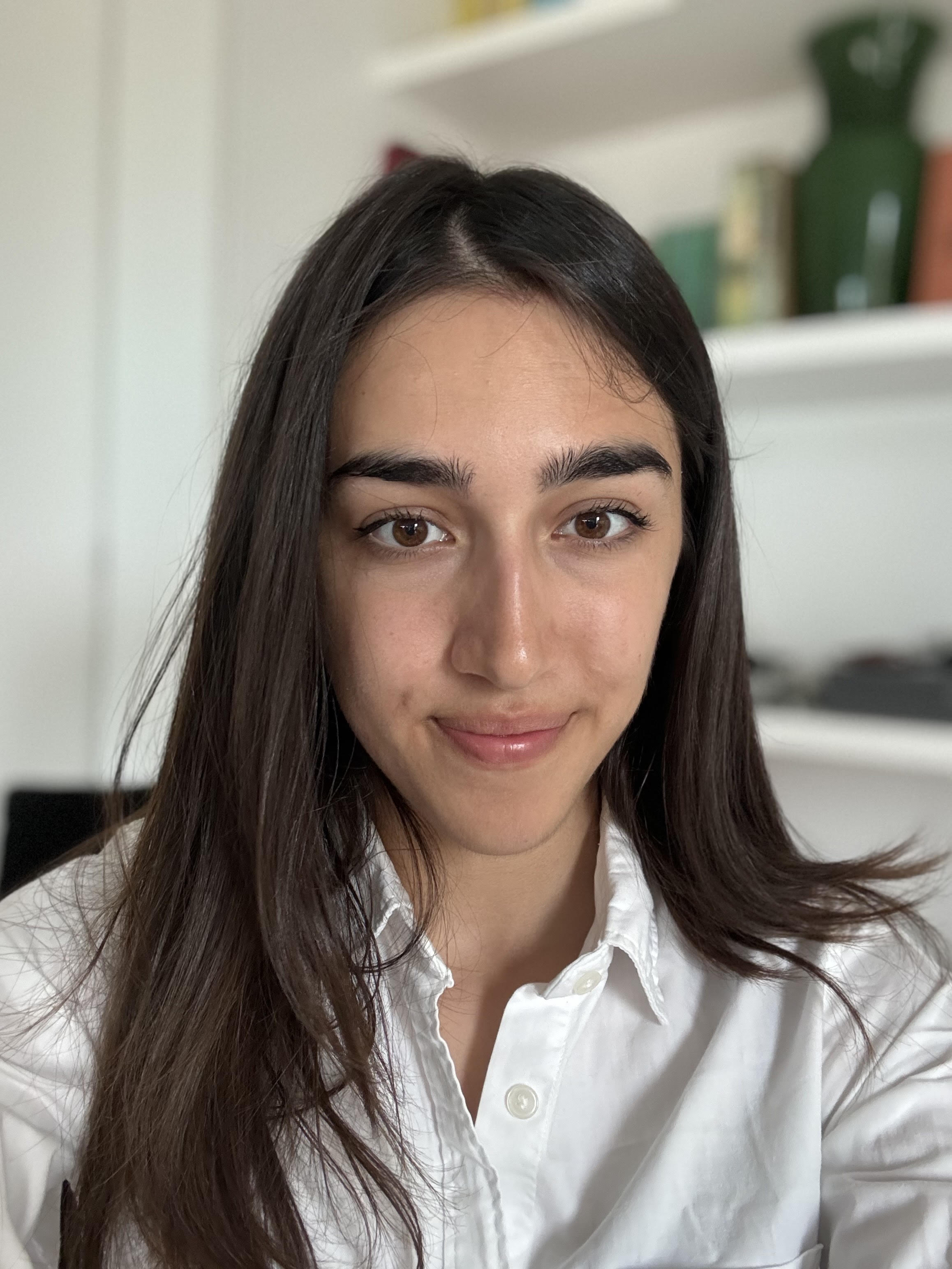 Portrait of a person with long dark hair in a white shirt, seated in front of a white bookshelf filled with books and decorative items, including a green vase.