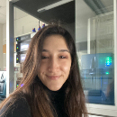 Selfie of a person in a professional workspace with electronic equipment and shelves in the background, suggesting a laboratory or technical office setting.