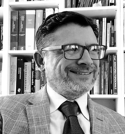 Black and white portrait of a smiling person in a suit and glasses, seated in front of a bookshelf filled with books.