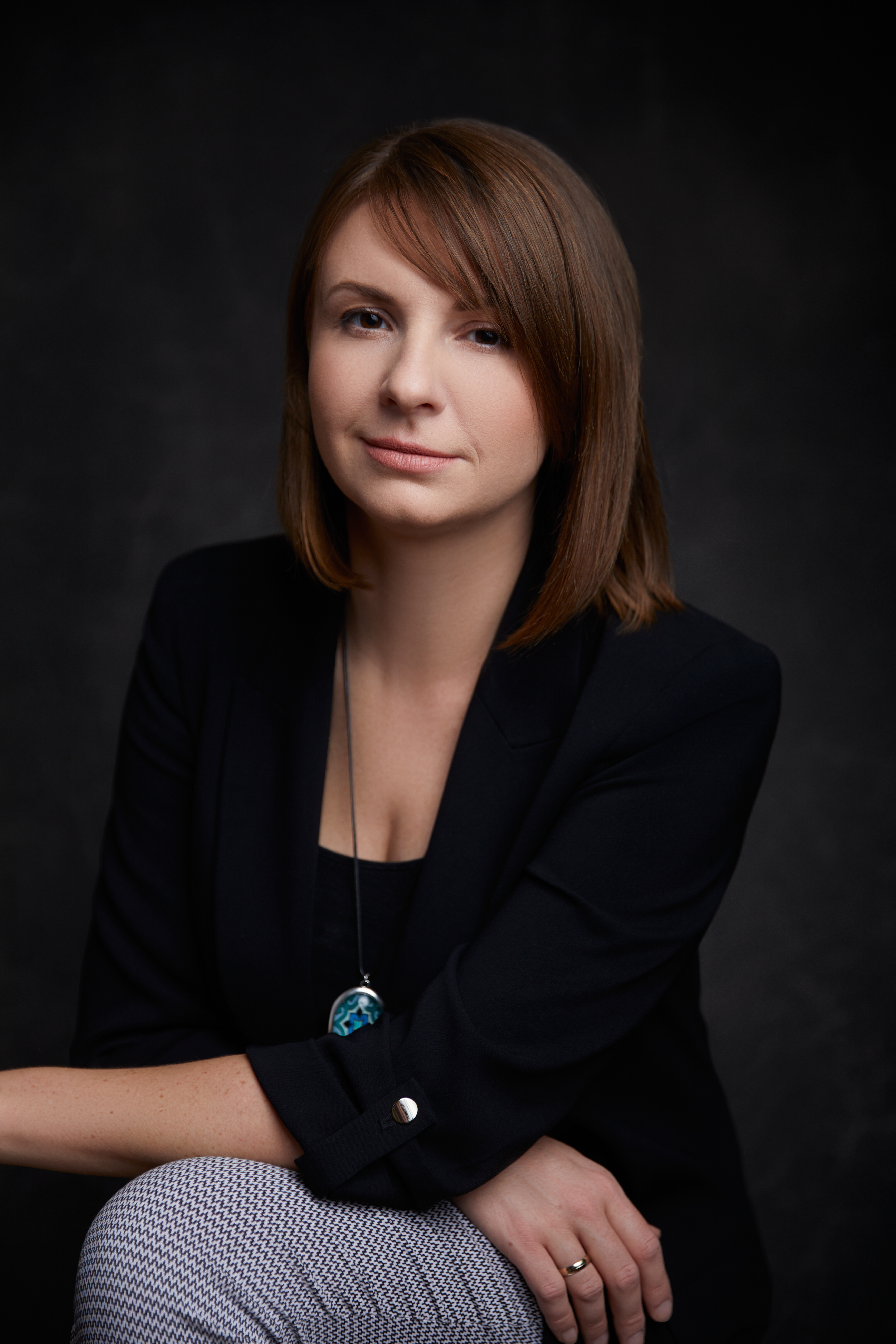 Professional portrait of a person in a dark blazer and patterned pants, seated against a dark studio background.